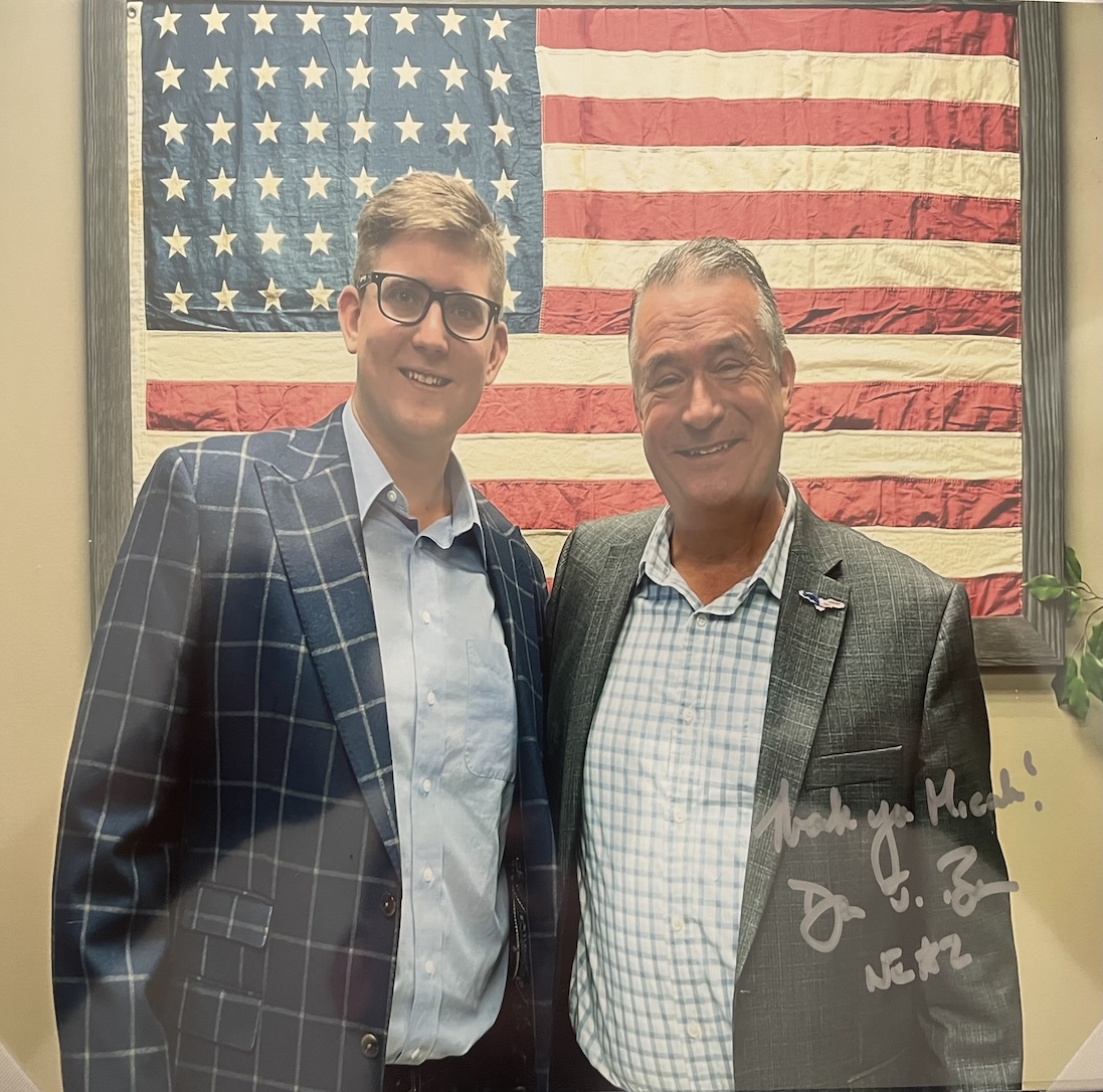 Micah, who is wearing a blue suit jacket and dark glasses, is pictured in front of the American flag with Congressman Don Bacon, who is wearing a gray suit jacket