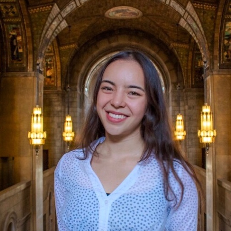 Smiling woman in white blouse, standing in a grand hall with ornate arches and warm lighting.