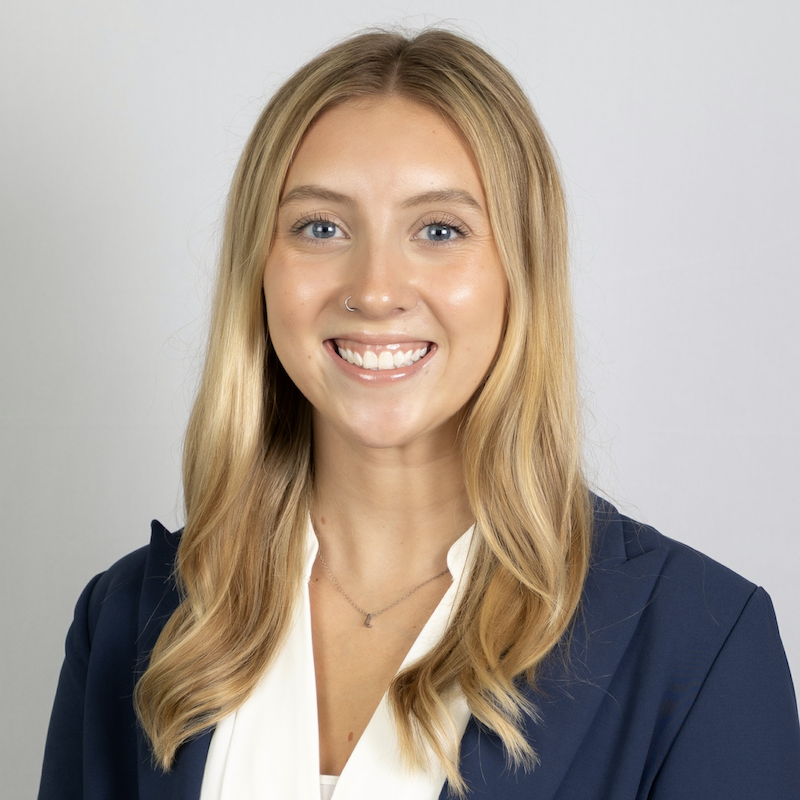 Smiling woman with long blonde hair wearing a navy blazer and white blouse against a gray background.