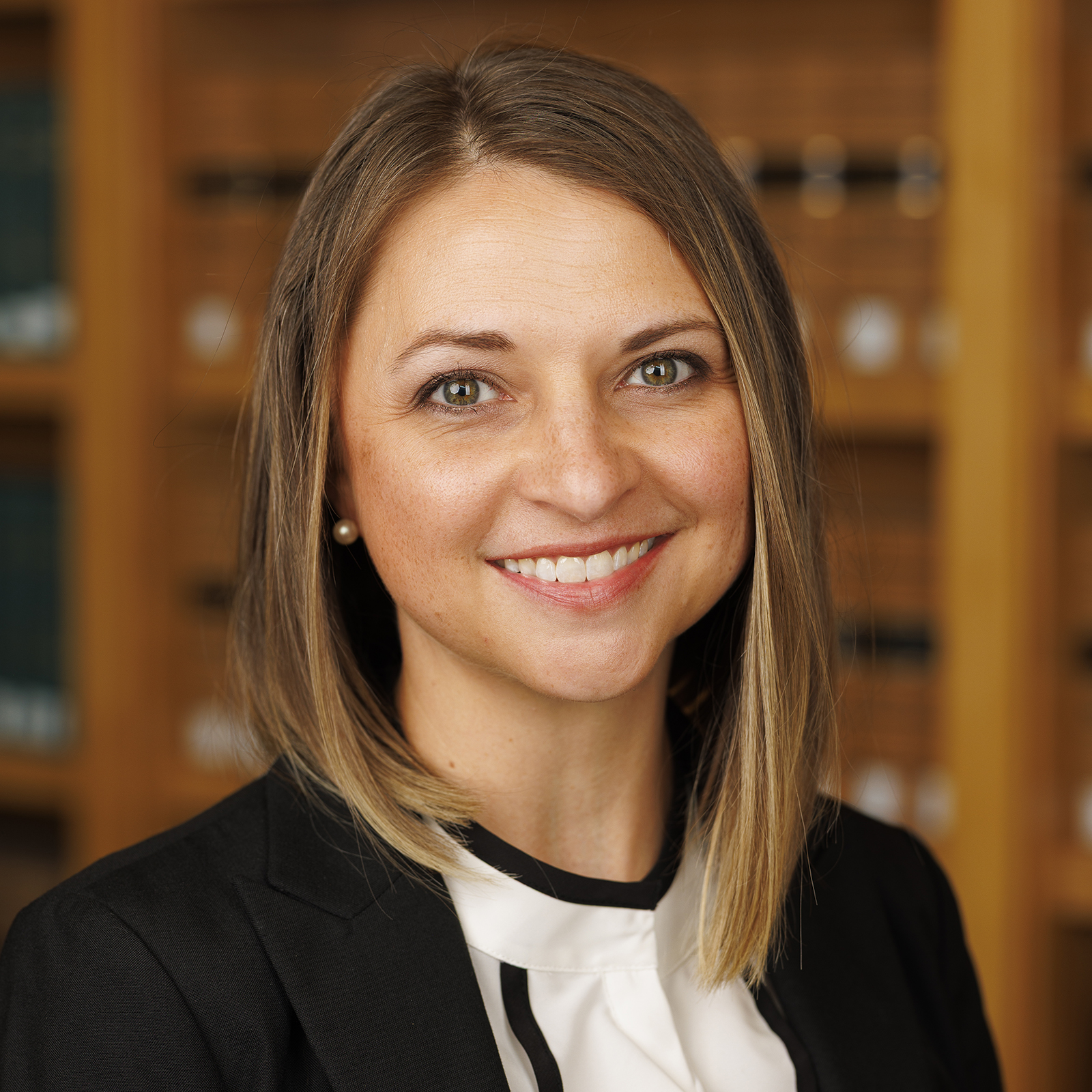 Smiling woman with shoulder-length hair, wearing a black blazer in a library setting.