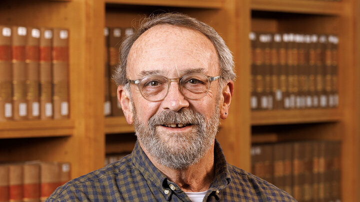 Richard Leiter, an older man with greying hair and a beard, stands in a brown button-down with glasses, in front of a bookcase.