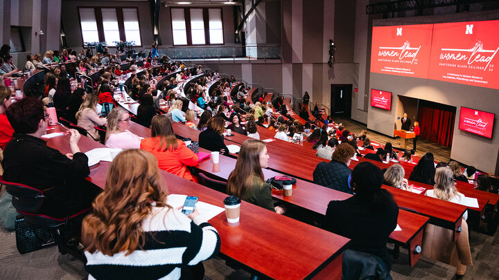 A crowd of people at red tables watch a woman present in front of a projector at a past Women Lead conference.