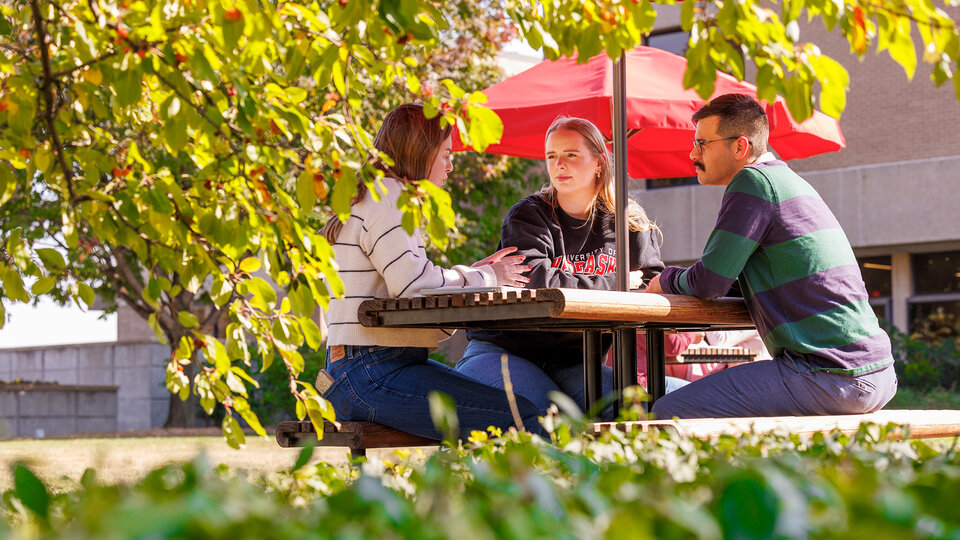 two female and one male students sitting at a picnic table under a red umbrella