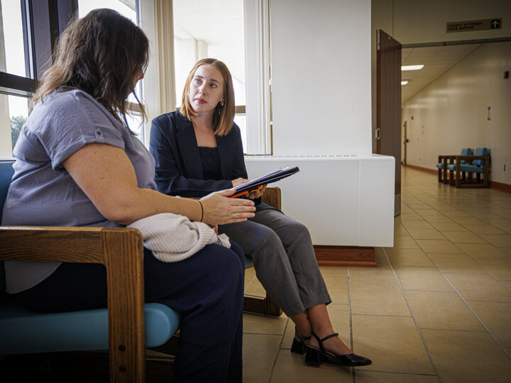 Two people sitting in a room with windows, one is holding a notepad while the other is speaking.