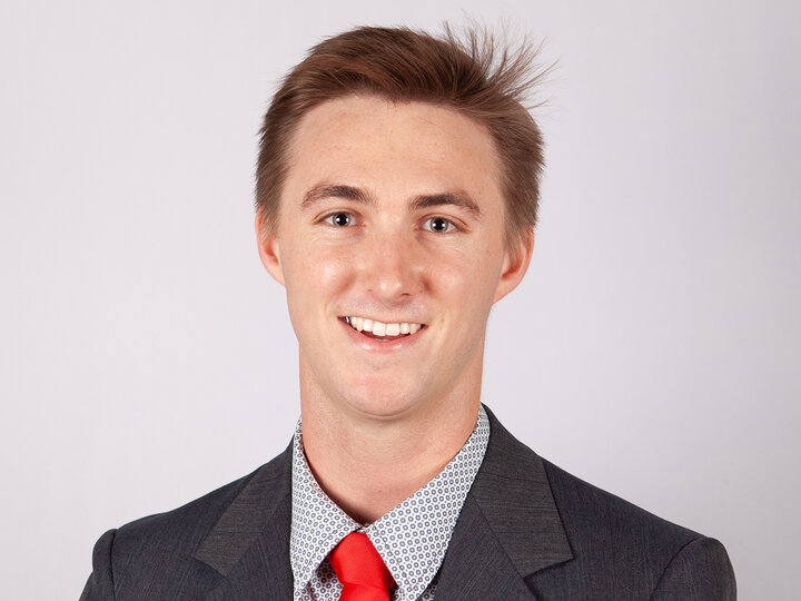Man smiling in a gray suit, red tie, and patterned shirt against a plain white background.