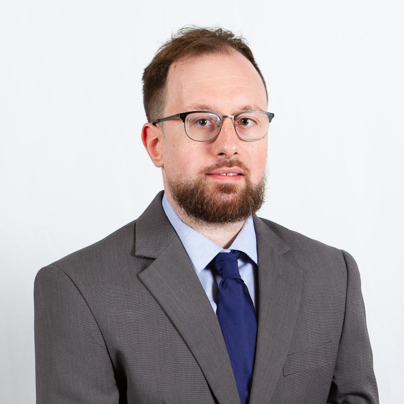 Portrait of Alan, who has short brown hair, glasses, and a beard and is wearing a suit and tie