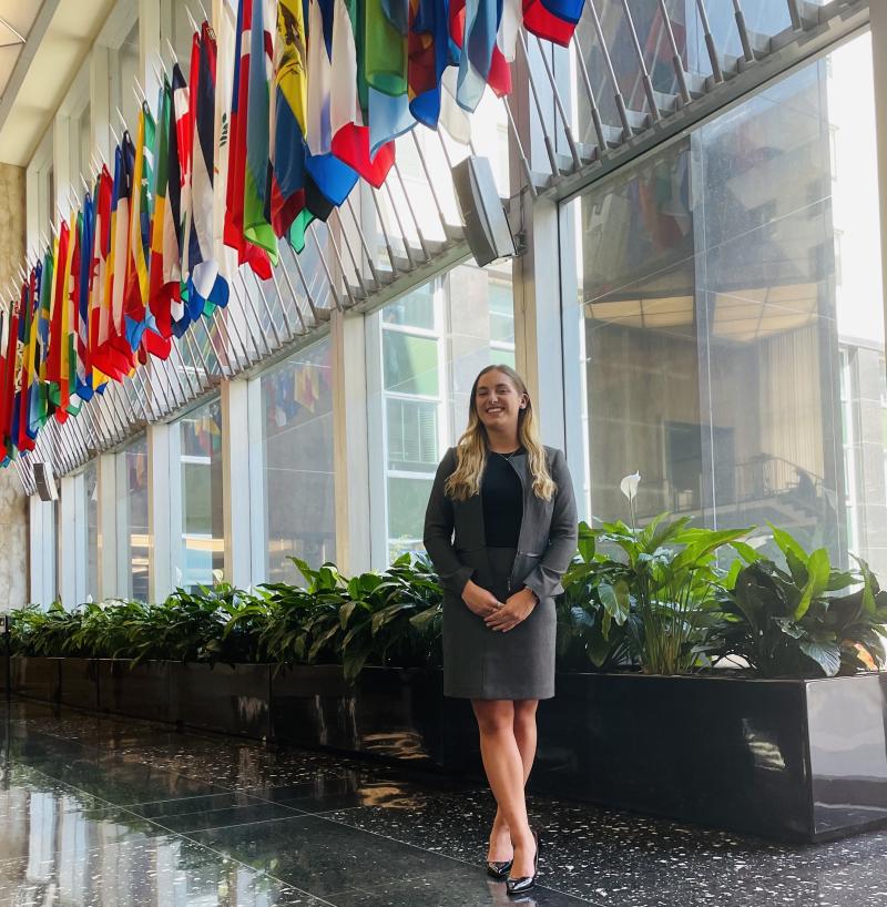 Emma is standing in front of wall of large windows and smiling at the camera. She has long blonde hair and is wearing a gray skirt suit. Above her is a long row of flags from different countries.