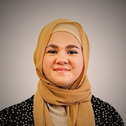 Jordan is pictured looking directly at the camera and smiling. She is wearing a black jacket and gold-colored hijab. 