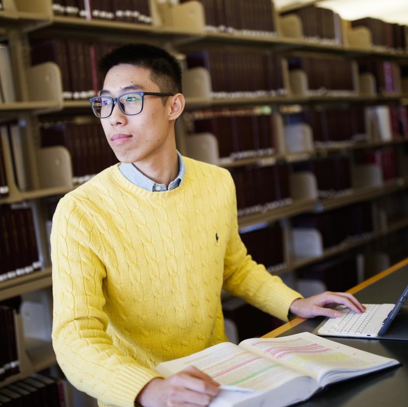 Kevin is standing in the library with a textbook on the table in front of him and is looking off to the side. He has tan skin and short, black hair and is wearing a yellow sweater and glasses. 