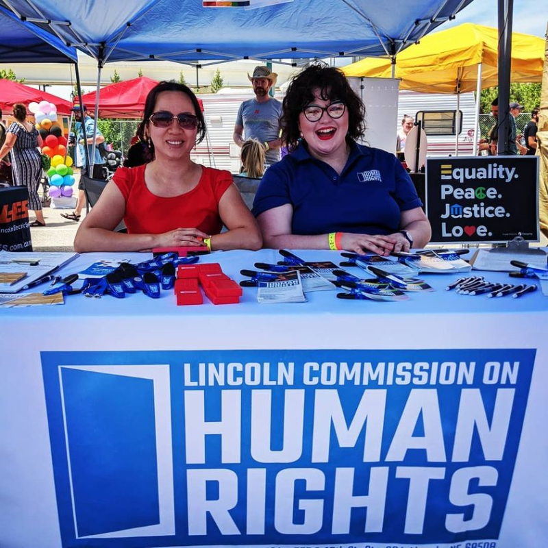 Two people are sitting at a table with a sign that says Lincoln Commission on Human Rights. They are looking at the camera and smiling. The person on the right is wearing a red shirt and sunglasses and has their hair tied back. Ivy is on the right wearing a dark blue polo shirt, eyeglasses, and red lipstick and has brown curly hair. 