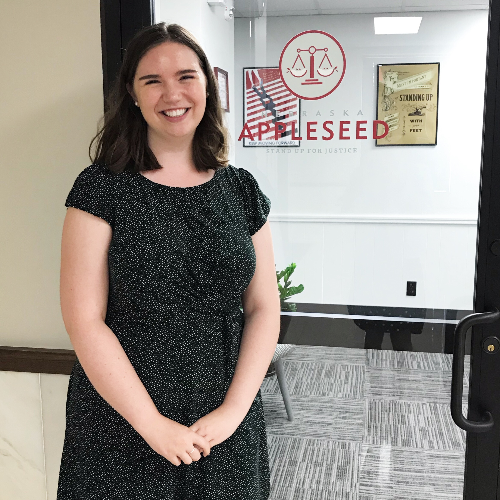 Claudia is wearing a black dress with short sleeves and is standing next to an office door with Nebraska Appleseed's name and logo. She has light skin, shoulder-length, straight brown hair, and is smiling at the camera. 