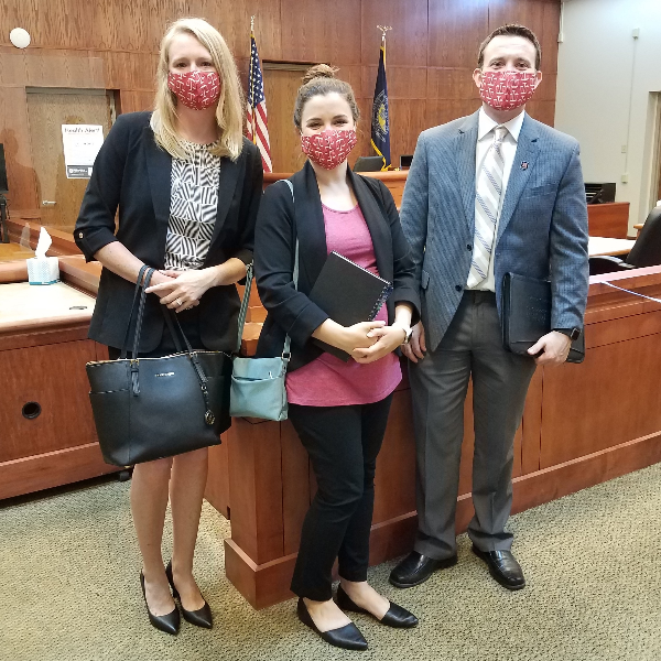 Three people standing in a courtroom are wearing face masks with the scales of justice and looking at the camera. 