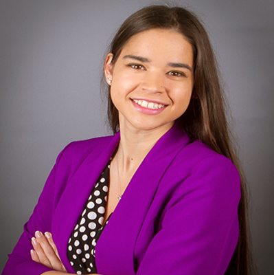 Shailana is pictured in a headshot with long brown hair and wearing a purple blazer. She is smiling at the camera. 